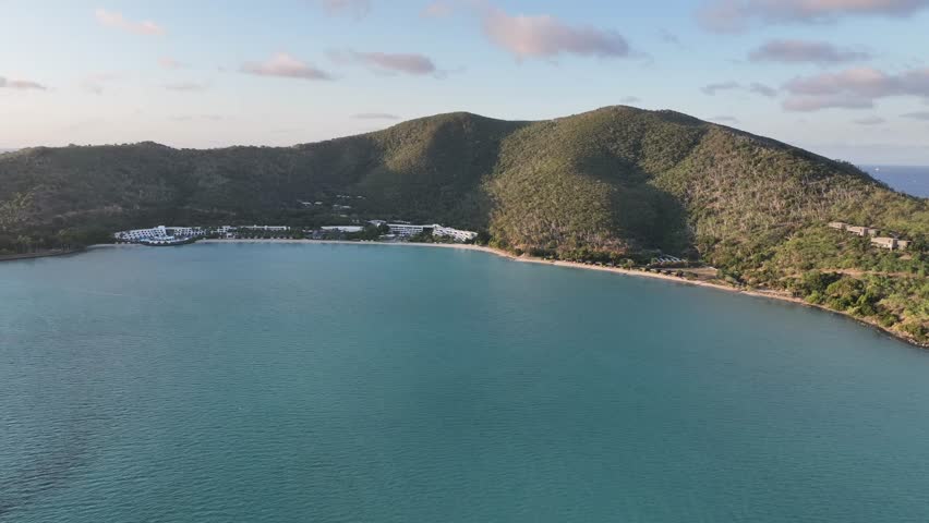 Wide drone shot at golden hour with a very slow pan right across Hayman Island’s sheltered bay, holding calm blue water, the beachfront resort and sunlit green hills, Whitsundays, Queensland, Australi