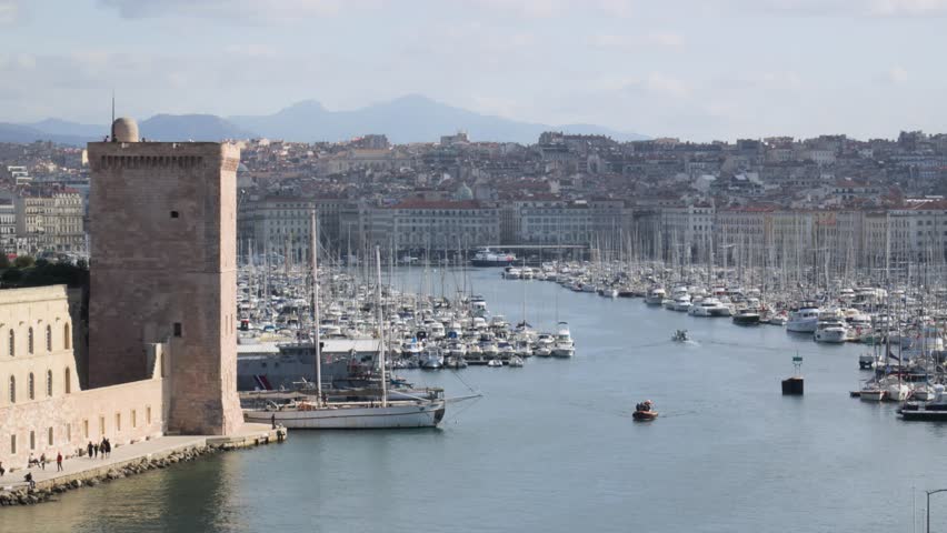 Telephoto shot from the sea toward Marseille harbor. Tour du Roi René stands out clearly, boats moving on the water, city skyline visible behind the port under a sunny cloudy sky.