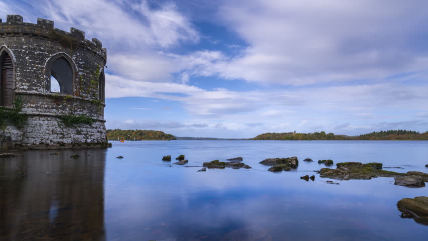 Timelapse of a medieval tower on lake with shoreline rocks in the foreground and medieval castle in the distance on a partially cloudy day in county Roscommon in Ireland.