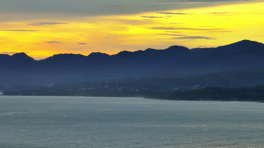 Panoramic view of mountain silhouettes and tranquil sea beneath vibrant golden sunset sky.