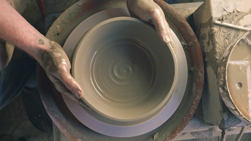 A craftsperson shapes a clay bowl on a pottery wheel, their hands skillfully guiding the wet clay into form. The workshop setting, with tools and clay remnants, highlights the art of pottery.