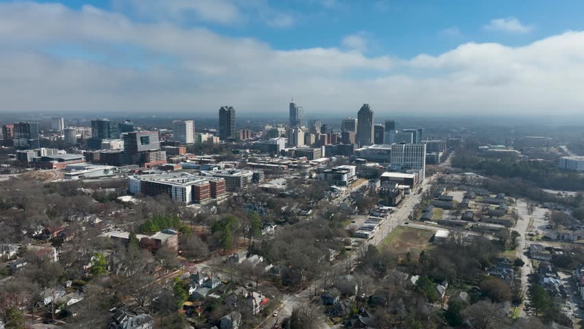 Aerial drone view of downtown Raleigh skyline with highway, residential neighborhoods, trees, and urban landscape under bright cloudy sky.