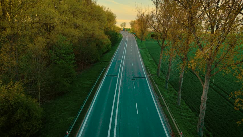 Multiple cars driving on open countryside road through fields and forest at sunset. Passenger vehicles moving along wide rural highway in warm evening light. Autos traveling across scenic landscape