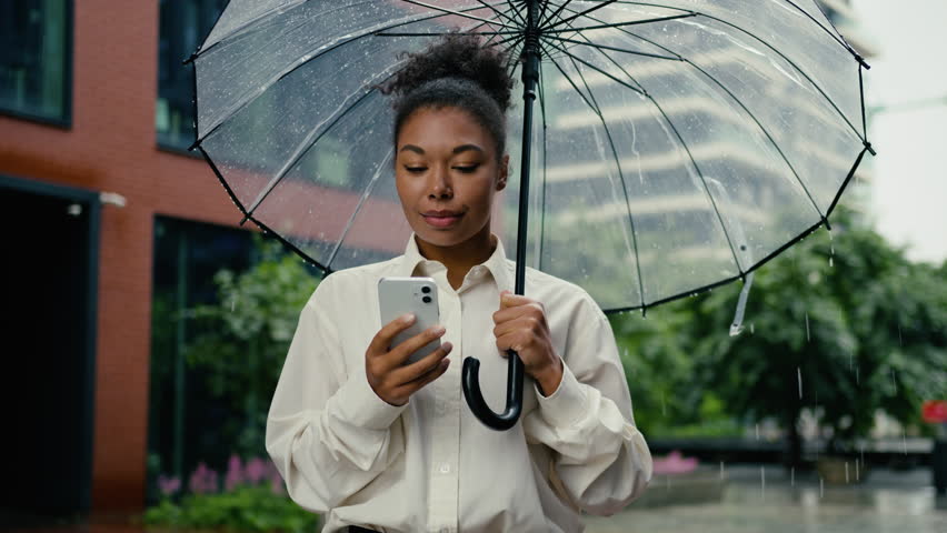 African American business woman with umbrella browsing mobile phone in rainy downtown smiling happy reading good news businesswoman female girl with smartphone enjoy using online app in rain weather