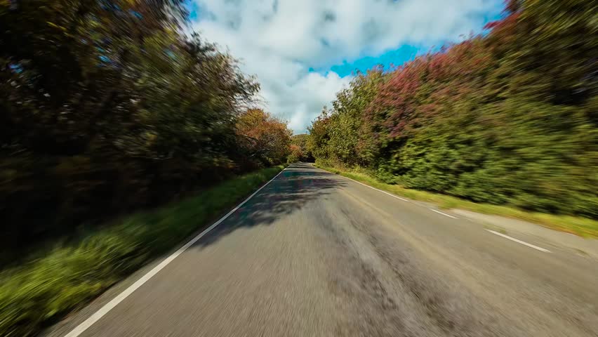 Driving along countryside road with left hand traffic through forested area in United Kingdom POV. Cruising down rural English lane surrounded by autumn trees and greenery first person view. Traveling