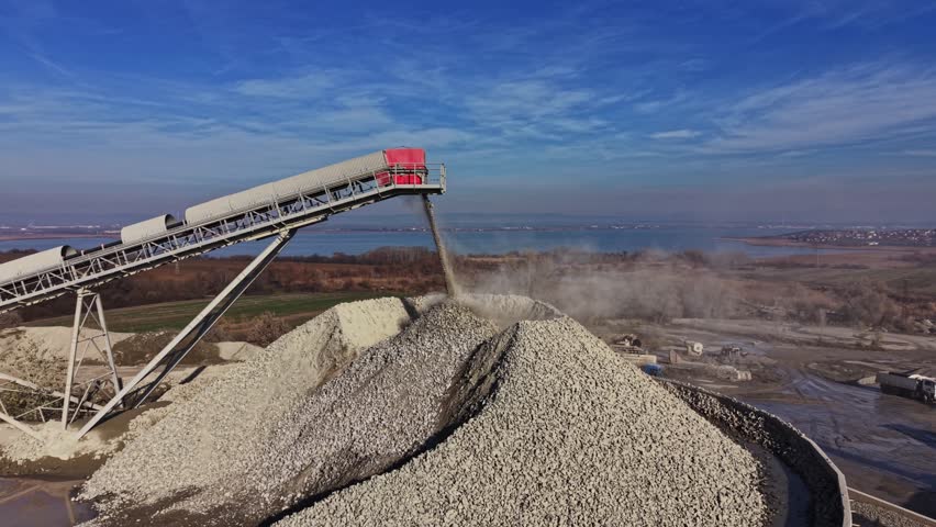 A conveyor belt transfers material at a quarry site. Large piles of rock and gravel are visible. The industrial activity takes place under a clear sky with a view of a water body.