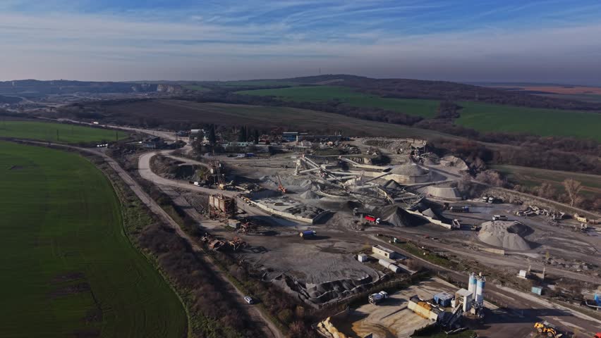 A quarry operation is underway in a rural setting. Heavy machinery moves materials while trucks are parked. Piles of stone and dirt are visible across the landscape under blue skies.