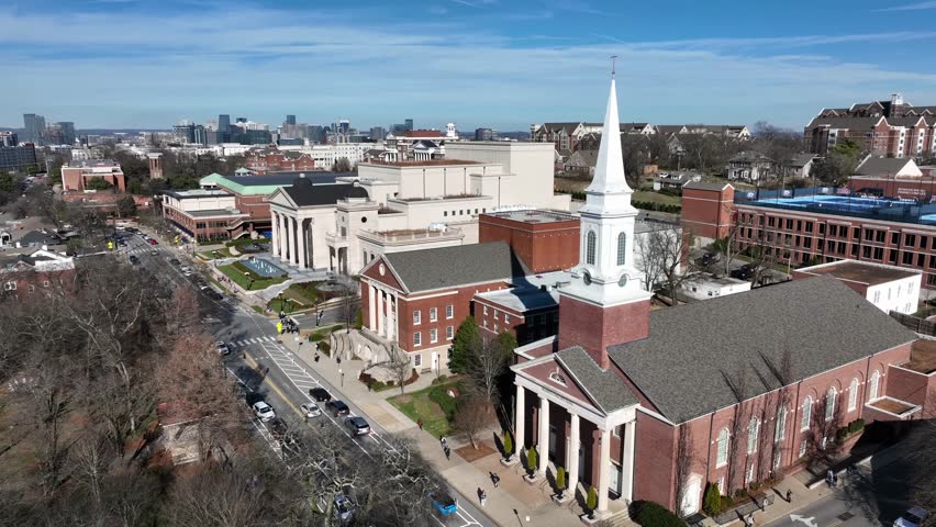 Rising aerial view of Belmont University in Nashville, Tennessee, showing red brick campus buildings and church steeple under clear blue sky with downtown skyline in distance.