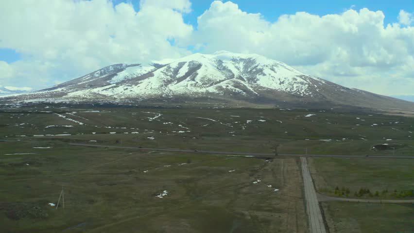 Mount Kazbek snow-covered peak rising above green plain in Caucasus Georgia, drone view highway, rural buildings, dramatic mountain landscape under cloudy sky.