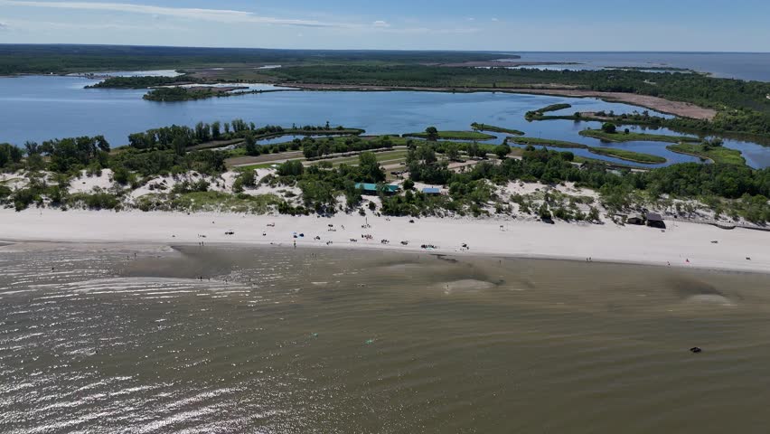Aerial from far over Lake Winnipeg looking east toward Grand Beach, Manitoba on a sunny summer day. The camera rotates right, revealing the sandy shoreline, beachgoers, lagoon, and boreal forest.