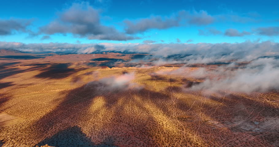 Flying in the feathery clouds floating over the deserted landscape. Numerous little shrubs cover the territory of Joshua Tree National Park, California, USA.