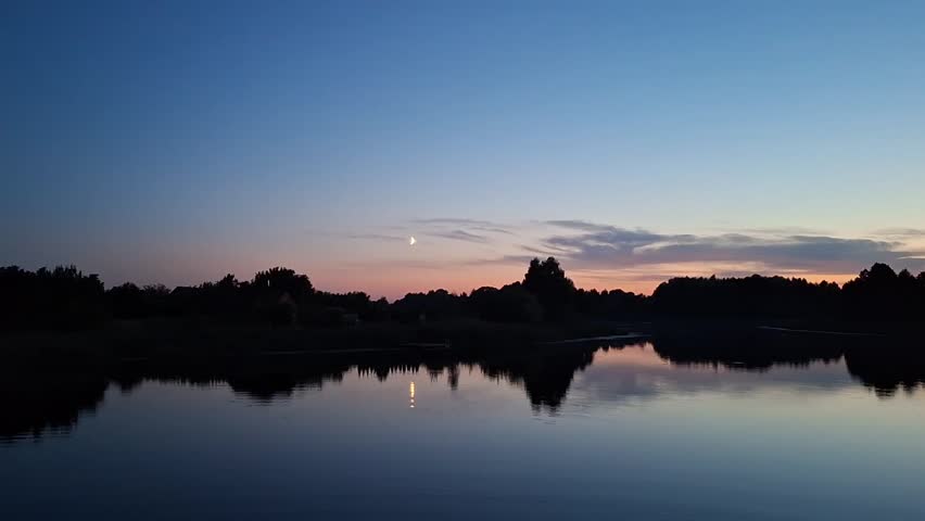 Calm summer sunset over the lake with a crescent moon reflecting in the water