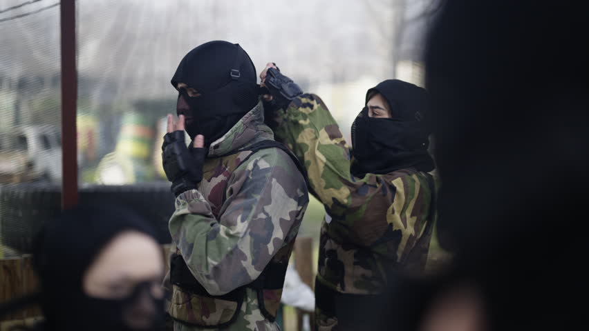 Participants in camouflage gear and protective masks prepare for an action sport. A team gets ready for a paintball or air soft game in an outdoor arena.