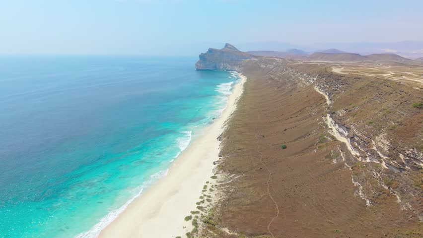 Aerial view of the turquoise ocean waves meeting a white sand beach beneath high desert cliffs on Socotra Island, Yemen.