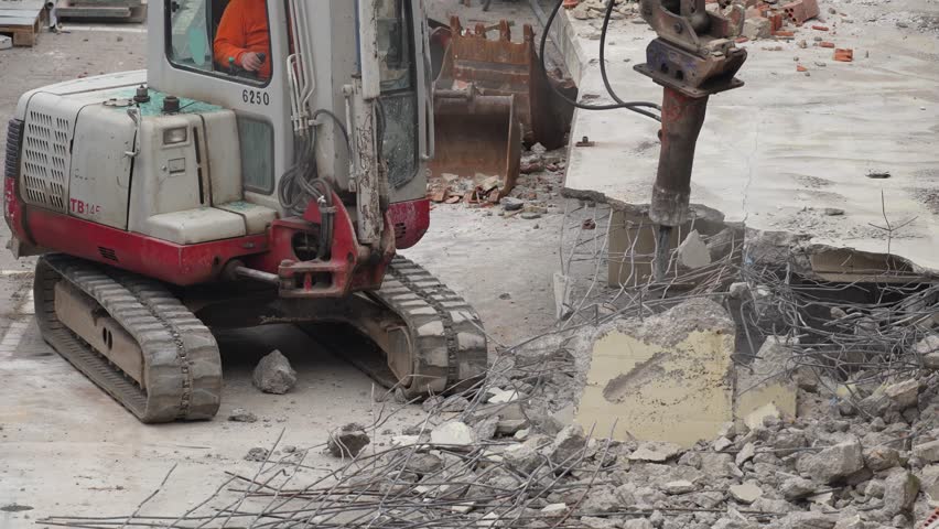 Wide demolition view with a compact excavator and hydraulic breaker breaking a concrete slab edge.
