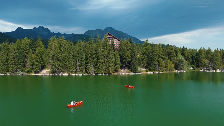 Drone view of rowboats on Štrbské Pleso in Slovakia with High Tatras mountains, forest shoreline and alpine hotel under dramatic sky.