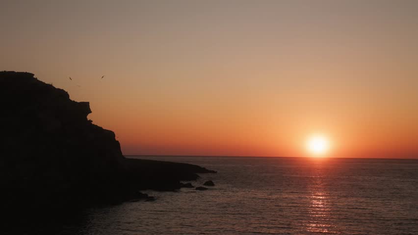 Static shot of sunset over Portinatx bay in Ibiza with golden reflection on calm Mediterranean sea and cliff silhouette.