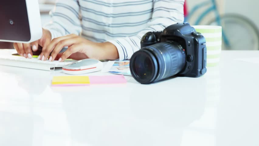 Pro typing at desk, reaching for DSLR, inspecting controls for shot while network icons overlaying. Camera, photography, studio, coworking, workspace, creative, bright