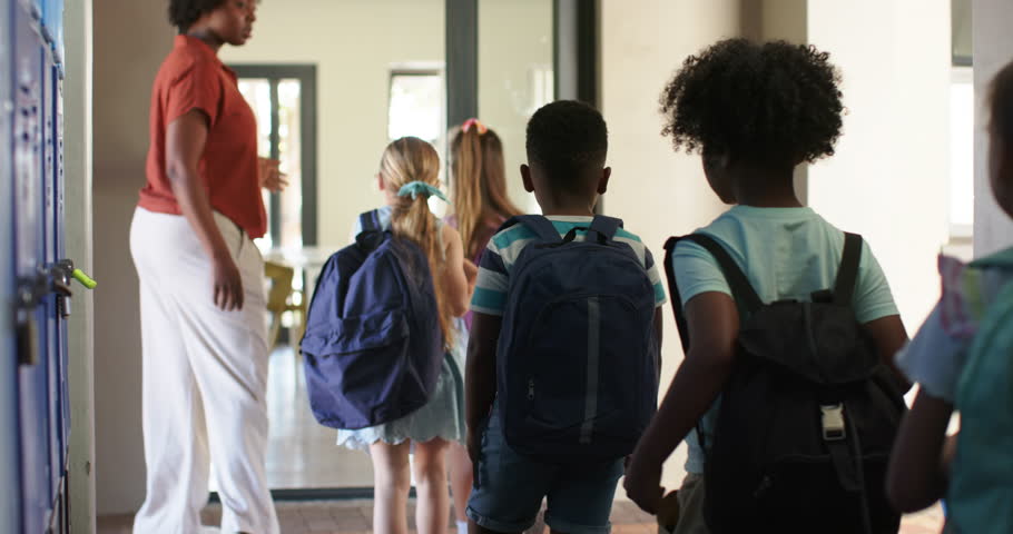 African American teacher gesturing, guiding children down school hall into doorway with backpacks. Educator, students, corridor, lockers, backpack, daylight, lineup