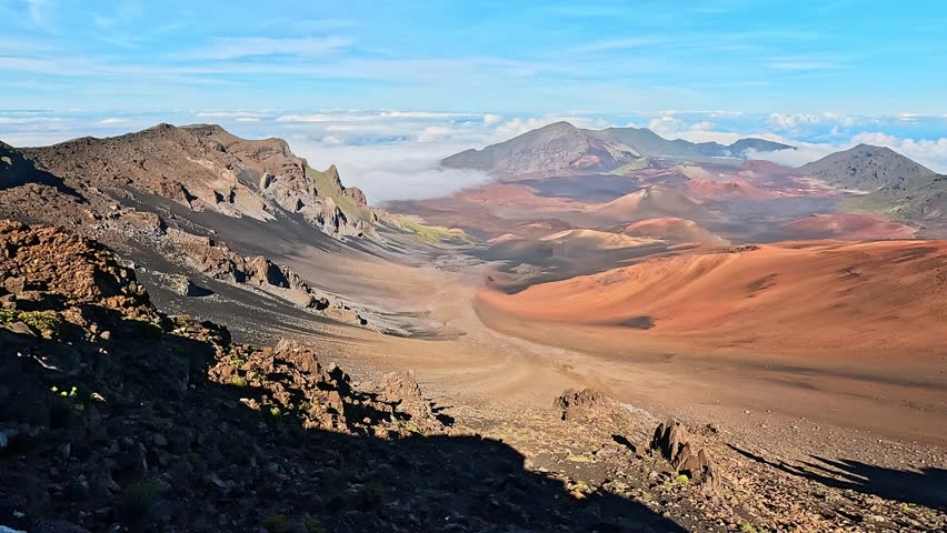 Cinematic 4K panning view of Haleakalā Crater on the island of Maui, Hawaii, filmed from the summit visitor center. The sweeping shot reveals the vast volcanic landscape filled with colorful cinder cones, rugged lava formations, and dramatic terrain stretching across the crater floor. A deep blue sky rises above a layer of clouds below the summit, emphasizing the high elevation and expansive perspective.

Haleakala is one of Hawaii’s most iconic natural landmarks, known for its surreal, Mars-like scenery and vibrant mineral tones ranging from red and brown to gold and gray. The panoramic motion highlights the scale and geological beauty of this dormant volcano.

Ideal for travel marketing, tourism campaigns, environmental content, geology themes, adventure storytelling, national park visuals, and destination branding. The dramatic volcanic textures and high-altitude cloud layer create a powerful and inspiring natural scene.

Shot in ultra-high definition 4K resolution with rich natural color and smooth cinematic movement, suitable for commercial or editorial productions.
