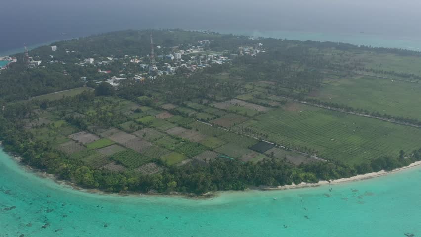 Wide aerial panoramic view of entire Thoddoo island revealing extensive agricultural farming fields, the local village, lush palm forests, and surrounding turquoise coral reefs.