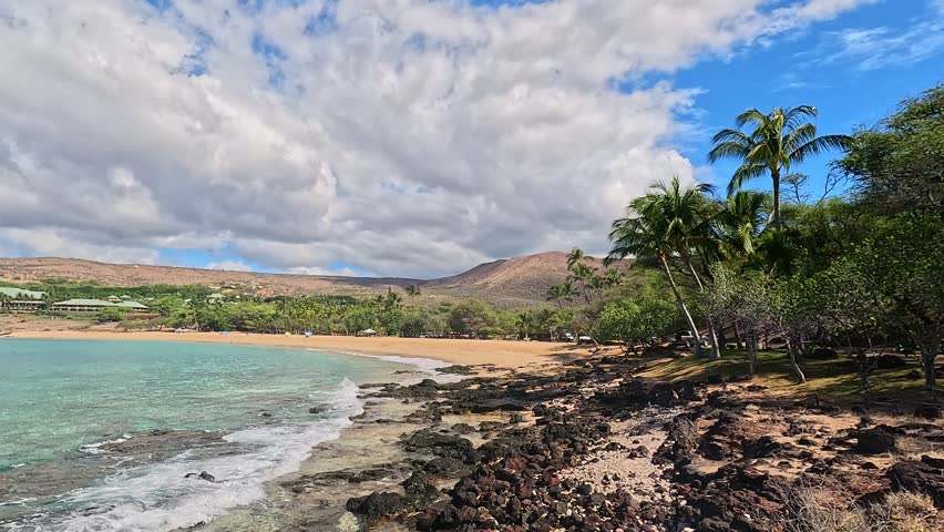 Cinematic 4K footage from one end of Hulopoe Beach on the island of Lanai, Hawaii, looking toward the luxurious Four Seasons Resort Lanai in the distance. The scene captures vibrant turquoise Pacific waters meeting warm golden sand as gentle waves roll onto the shoreline. A small reef formation is visible in the foreground, adding natural texture and depth to the composition.

A few visitors relax under umbrellas along the beach, enhancing the sense of scale while maintaining a quiet, peaceful, and remote atmosphere. Palm trees frame the tropical coastline, reinforcing the feeling of exclusivity and island escape.

This footage conveys the essence of a high-end Hawaiian destination — serene, uncrowded, and naturally beautiful. Ideal for travel marketing, luxury resort promotion, vacation branding, lifestyle content, wellness themes, tourism campaigns, and tropical destination storytelling. The calm ocean movement and expansive beach setting create a relaxing, inviting mood perfect for commercial or editorial use.

Shot in ultra-high definition 4K resolution, delivering rich natural color, fine sand texture, detailed reef elements, and cinematic coastal quality suitable for professional productions.