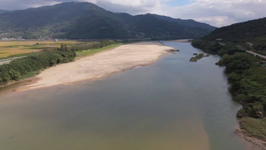An aerial perspective shows a wide river with a prominent sandy shoal stretching across the water, bordered by lush green forests and hills. In the distant background, rolling mountains rise above agricultural fields and small villages under an overcast sky. Concept of serene natural beauty