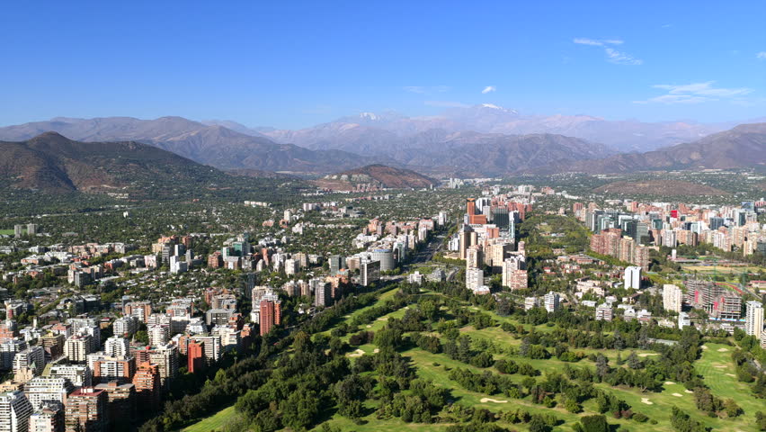 Vitacura neighborhood in Santiago Metropolitan Region seen from above with Parque Bicentenario green area and Andes mountain range forming dramatic backdrop, drone slow glide.