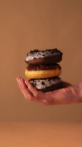 Close up of hands stacking the final vanilla glazed donut with colorful sprinkles on top of chocolate iced and cookie crumb donuts. Warm beige studio background with clean minimal look.
