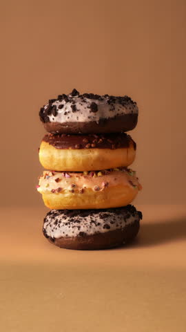 Hand removes donuts one by one from stacked tower with chocolate, cream and sprinkle toppings on warm beige background. Clean minimal setup ideal for ads, reels and food branding hook visuals.