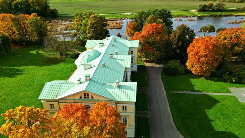 Historic manor with green roof beside river and vibrant autumn trees in European countryside
