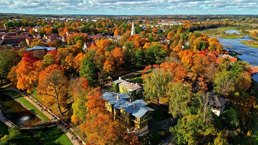 Drone panorama of European town with colorful autumn park, modern pavilion, church spire, and river.