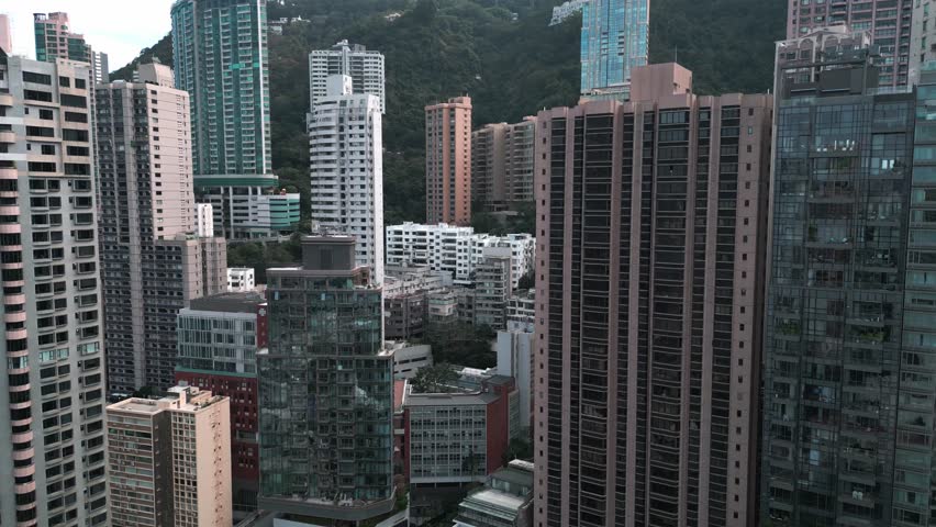 Aerial scenic drone footage of buildings and skyscrapers in downtown Hong Kong on a sunny day, highlighting the dense skyline and modern urban architecture.
