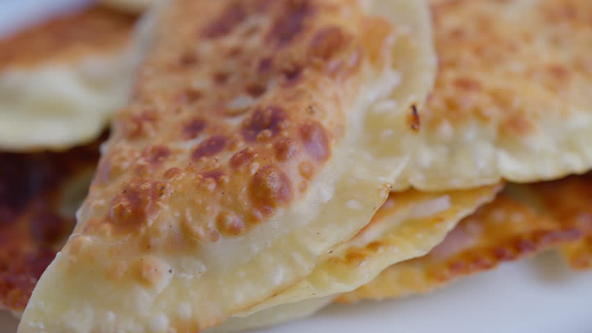 Golden fried chebureki on plate stacked closeup showing blistered browned crust flaky edges oil sheen savory meat filling hint rustic homemade presentation warm snack