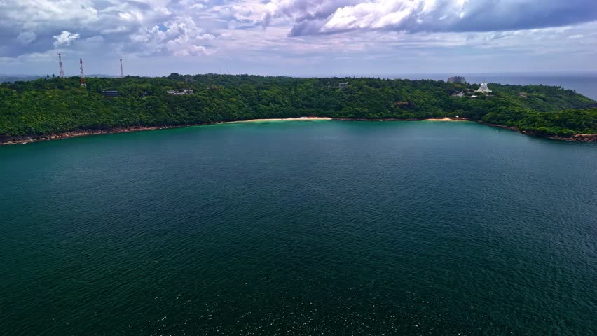 Wide aerial view of the Rumassala coastline with a calm bay, lush green hills, and cloudy sky, captured by a drone from high altitude.