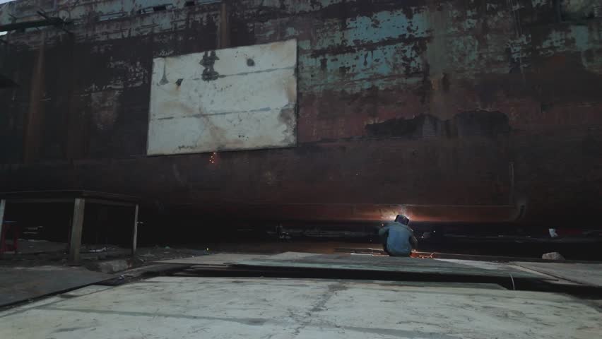 Wide shot of an industrial worker performing welding work on a massive rusted metal structure inside a shipyard or heavy manufacturing facility. Bright welding sparks illuminate the dark industrial environment, highlighting steel fabrication, metal repair, and shipbuilding processes. Perfect for themes related to heavy industry, engineering, construction, labor workforce, and industrial production.