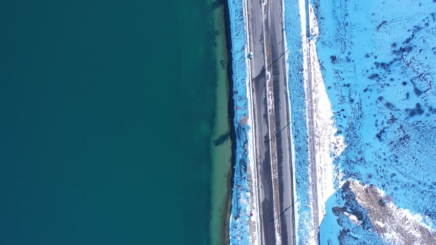 Straight highway between frozen snowy land and blue lake water, drone topdown view strong winter contrast, ice, road, nature patterns.