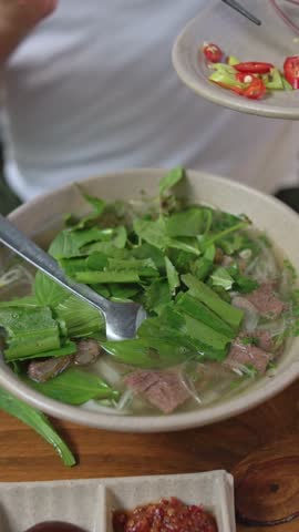 Vertical Screen Bowl Of Vietnamese Pho With Caucasian Diner Adding Sliced Chili Using Chopsticks, Vivid Red Peppers Against Green Herbs, Dipping Sauces On Side, Bold Spicy Vibe
