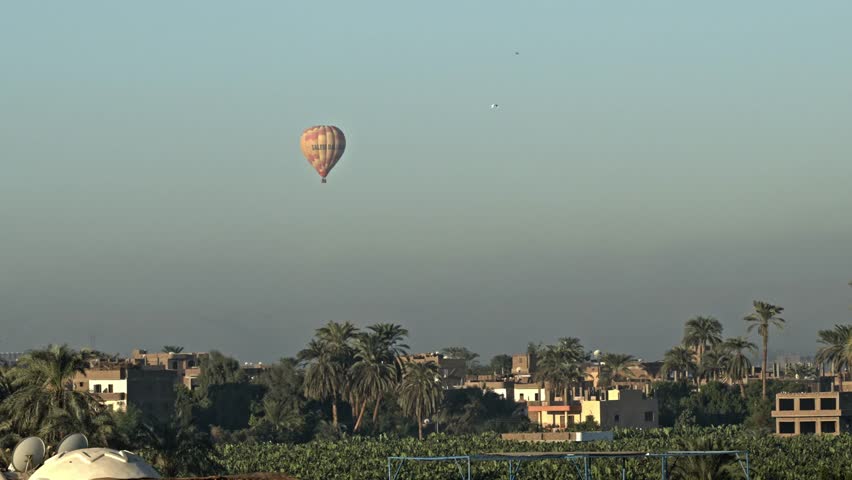 A stunning sunrise view over the West Bank of Luxor, Egypt. The image captures a brightly colored hot air balloon floating high above the rugged limestone cliffs of the Theban Necropolis.
