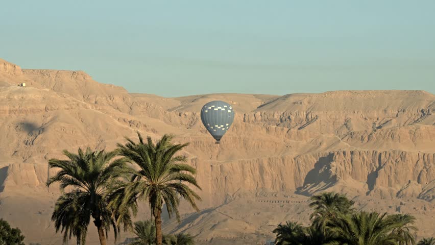 Sunrise over the Valley of the Kings in Luxor, Egypt. As the sun’s first rays hit the rugged limestone peaks of the Theban Mountains, a vibrant hot air balloon floats gracefully.