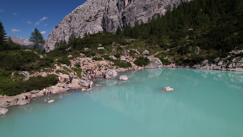 Aerial drone footage flying backwards above the turquoise waters of Lake Sorapis in the Dolomites, revealing the rocky shoreline and alpine landscape. The camera glides smoothly over the lake while distant mountain peaks slowly come into view, showcasing the pristine nature and dramatic scenery of this iconic alpine location.