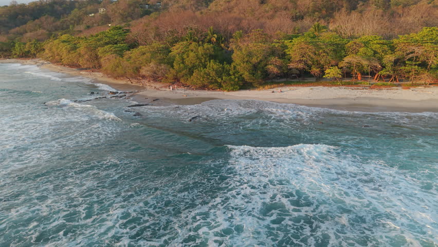 Smooth aerial reveal rising from ocean waves to tropical coastline and dense green vegetation in Santa Teresa Costa Rica