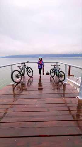 Vertical format of a young girl standing at the end of a boardwalk after the rain, along with two electric bikes, eating a snack. Thick clouds, mountain and body of water in the background.
