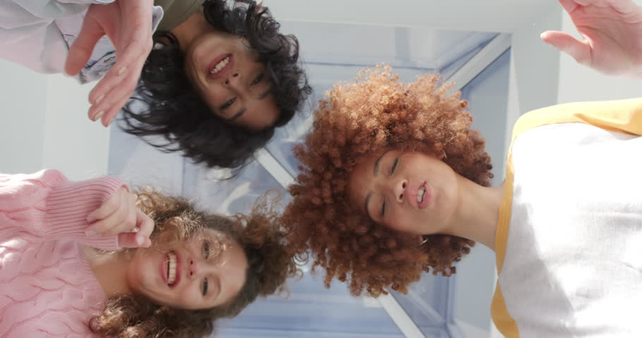 Diverse female friends leaning over device tapping to record, pointing and laughing in sunroom. Friendship, diversity, laughter, candid, casual, vibrant, social