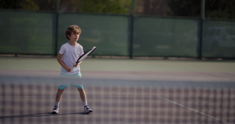 A young curly-haired boy swings his tennis racket at a yellow ball on an outdoor tennis court, demonstrating focus and skill in the sport.