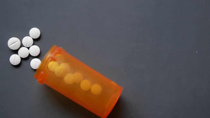 A motion footage of white pills spilling out of an orange prescription bottle onto a dark gray surface, yellow pills remaining inside