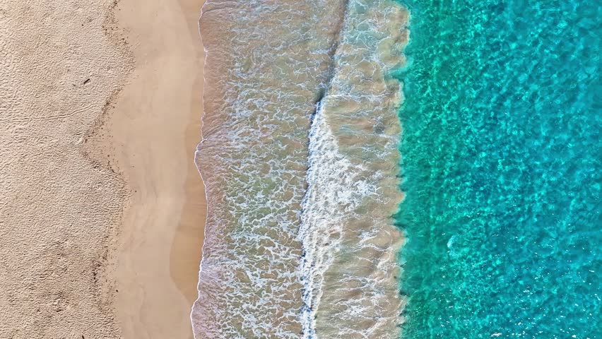 A sandy sea beach background with turquoise sea waves. Looping ocean texture, aerial view of calmly moving ocean waves. Flying over a sandy beach and waves, seamless beach loop.