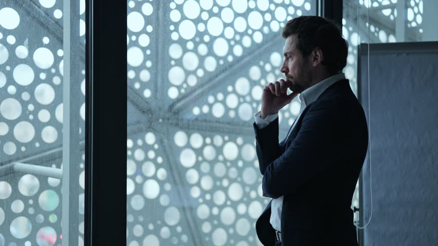 Businessman looks out window in deep thought. Contemplation and quiet reflection fill the moment. Pondering future, challenges, and solutions, expressing determination and focus in corporate setting.