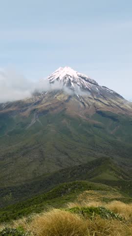 Timelapse of Mount Taranaki’s snow-capped peak as clouds drift across the summit above green volcanic slopes under a clear blue sky, scenic New Zealand landscape, vertical.