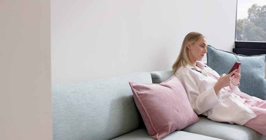 Adult woman holding red phone, scrolling, placing it, leaning back and relaxing on sofa, copy space. Pastel, minimalist, daylight, window, cushions, plant, calm